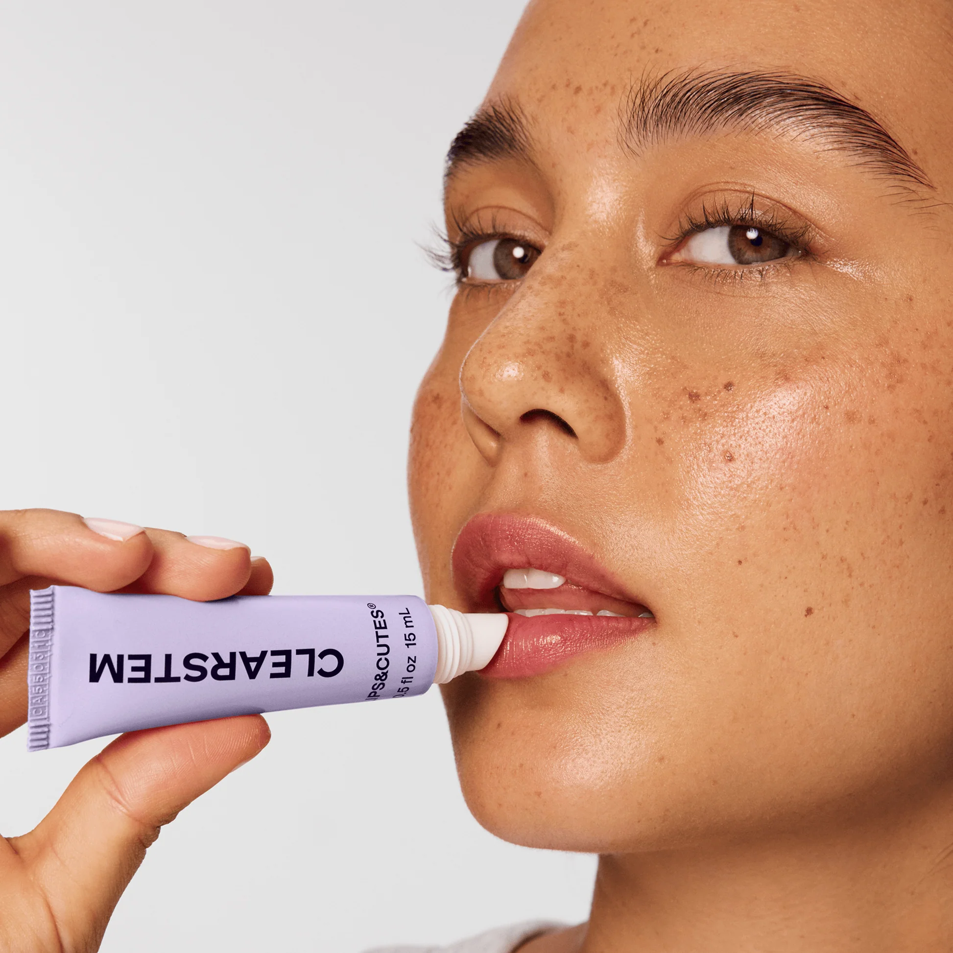 A person with freckles applies CLEARSTEM lip treatment from a lavender tube to their lips.