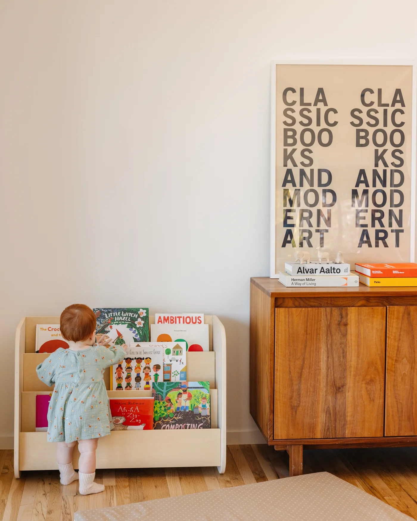 A toddler in a light blue dress stands at a small bookshelf, reaching for books. A wooden cabinet and framed art are on the right.
