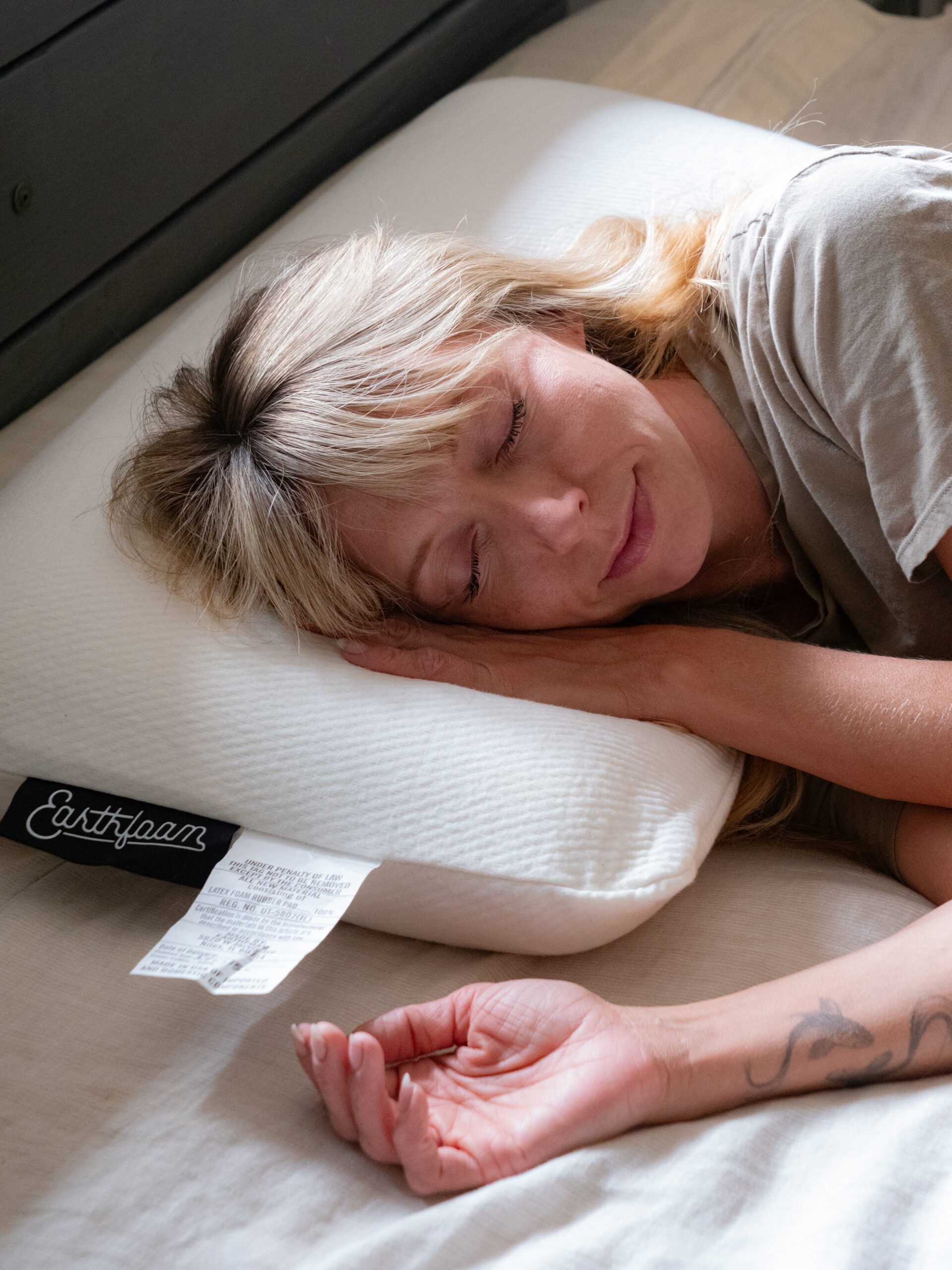 A woman with blonde hair sleeps on a white pillow labeled "Earthfoam" on a bed, resting her head and arm comfortably.