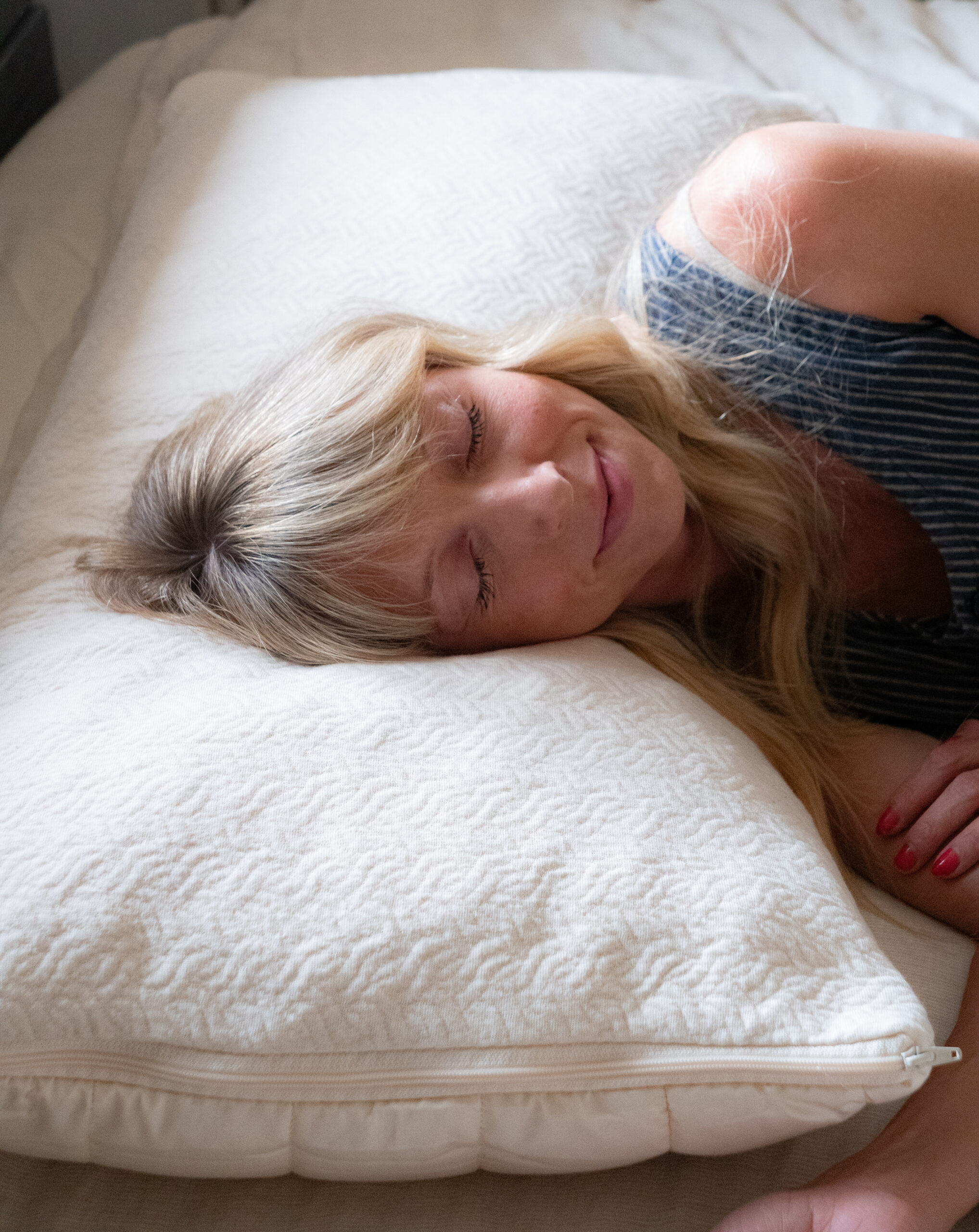 Woman with blonde hair lies on her side, smiling with eyes closed, resting on a large white pillow on a bed.