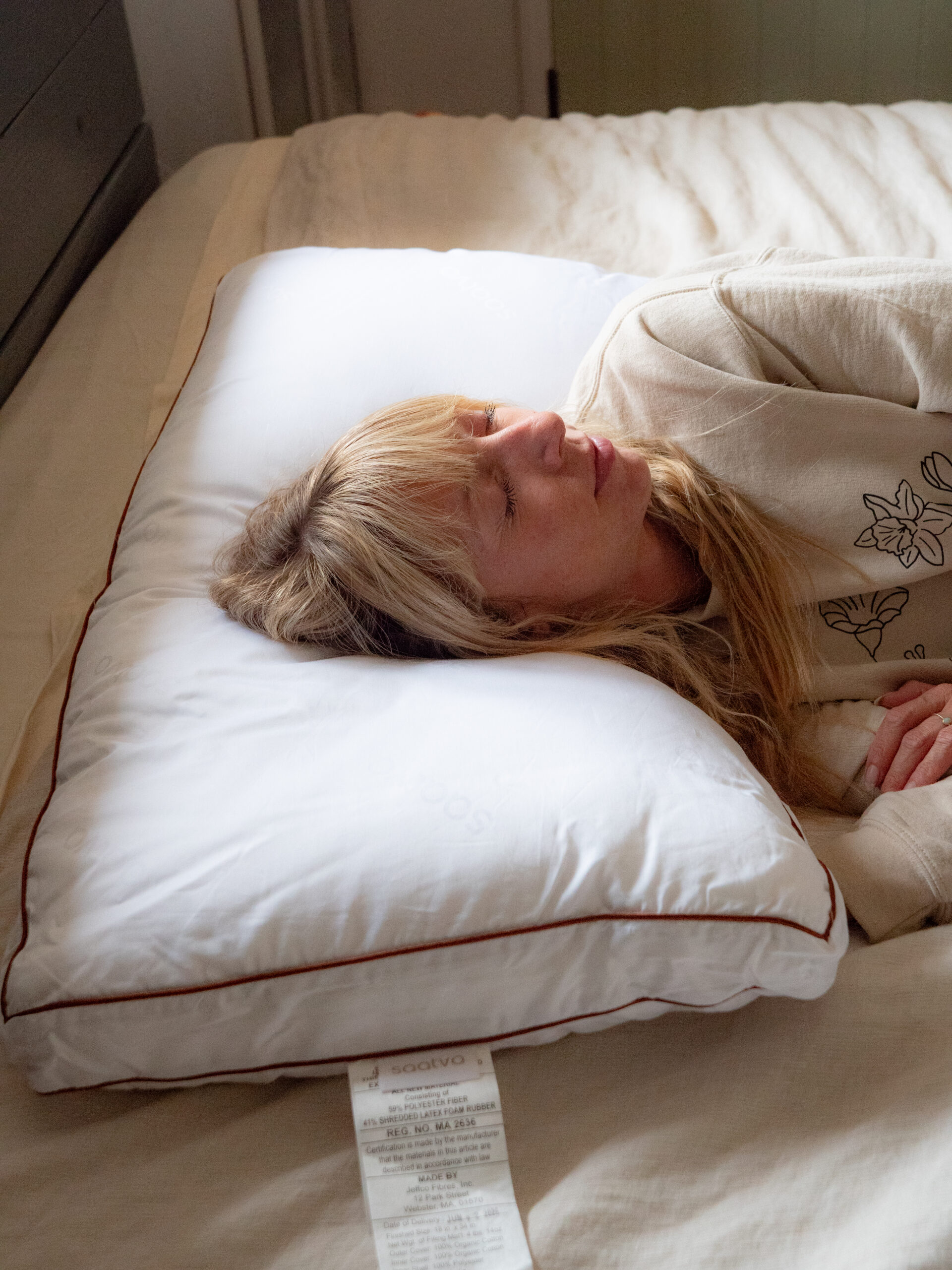 Woman lying on her side on a bed, resting her head on a large white pillow with a visible tag hanging from the edge.