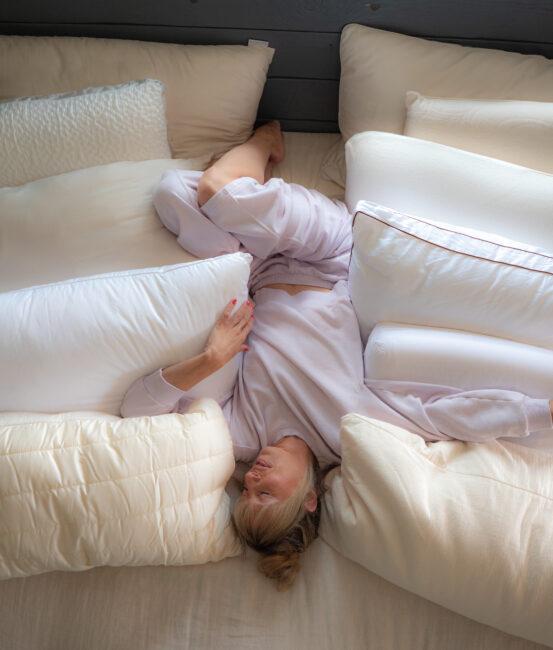 A person in light pajamas is lying in bed surrounded by multiple large white pillows, with one leg bent and arm resting on a pillow.