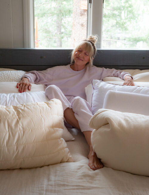 A woman in light loungewear sits relaxed on a bed surrounded by pillows, smiling with her eyes closed. Natural light comes through a window behind her.
