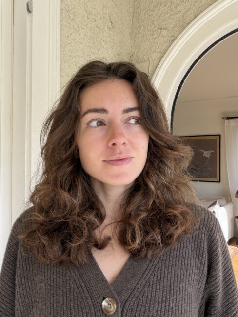 Woman with wavy brown hair wearing a brown sweater stands indoors, looking slightly to the right. A framed picture and arched doorway are visible in the background.