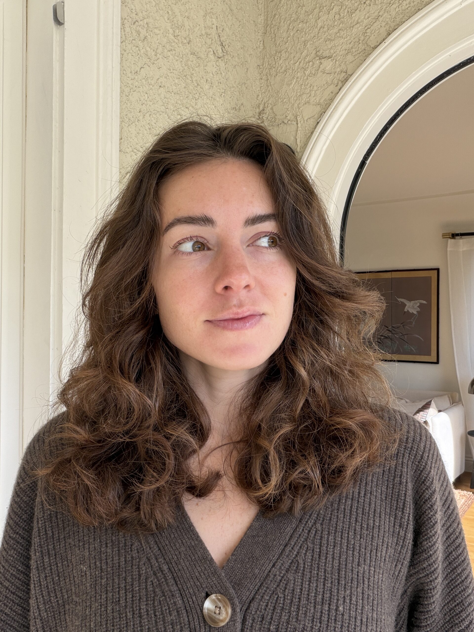 Woman with wavy brown hair wearing a brown sweater stands indoors, looking slightly to the right. A framed picture and arched doorway are visible in the background.