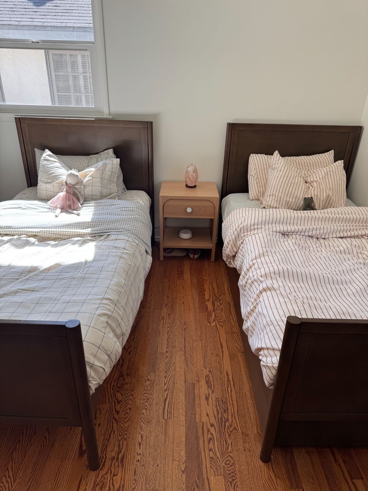 Two twin beds with striped bedding are separated by a small wooden nightstand with a lamp, set on a hardwood floor under a window.