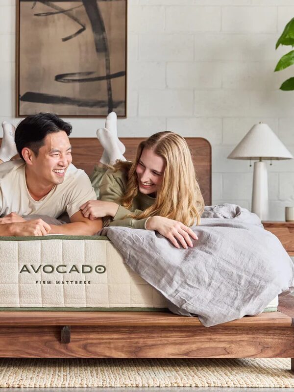 Two people lie on a bed with an Avocado firm mattress in a modern, light-filled bedroom with wooden furniture and plants.