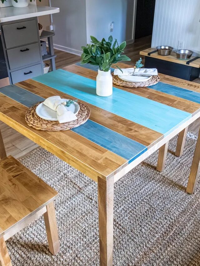 A wooden dining table with blue painted stripes, set for two with woven placemats, napkins, and a vase of greenery, stands on a woven rug in a kitchen.