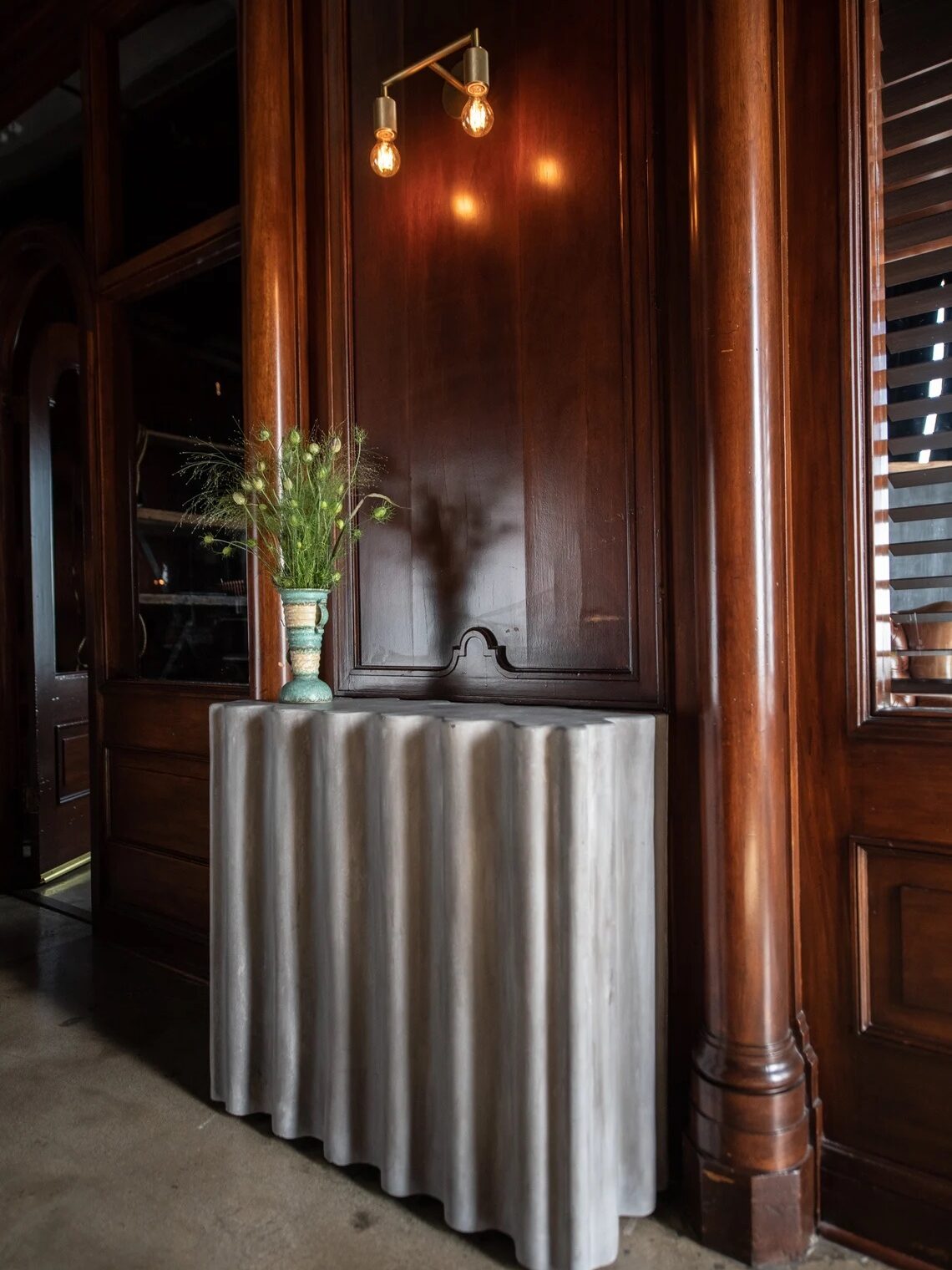 A gray, wavy-textured console table with a vase of flowers stands against a dark wood-paneled wall beneath a modern wall sconce with two exposed bulbs.