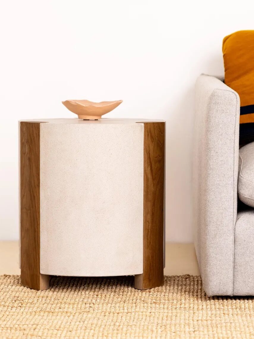 A small side table with wooden legs and a light-colored cylindrical body sits next to a light gray sofa with a mustard yellow and black blanket. A ceramic dish is on the table.