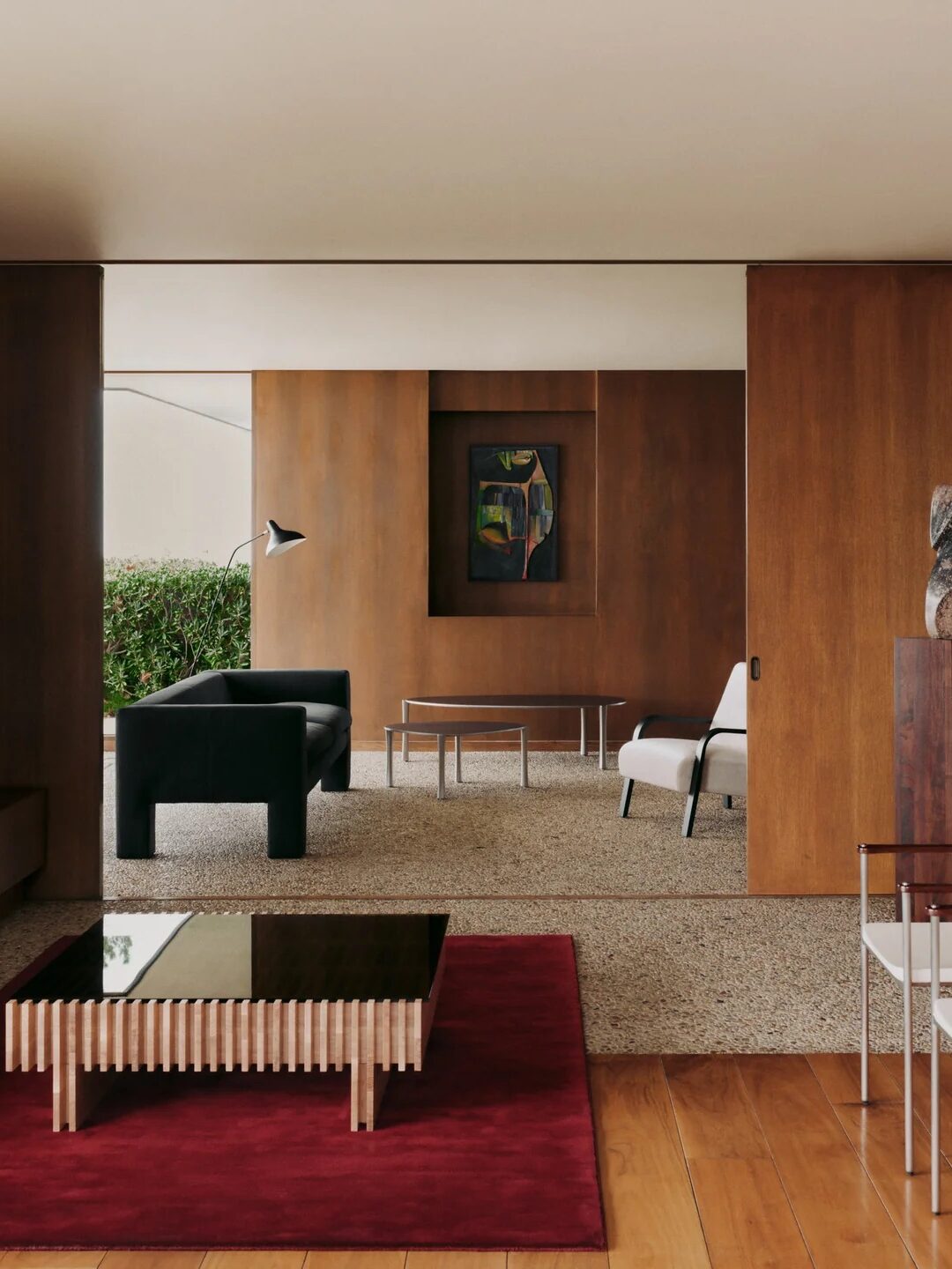 Modern living room with wood-paneled walls, minimalist furniture, a black sofa, glass coffee table on a red rug, and artwork on the wall. Large window lets in natural light.