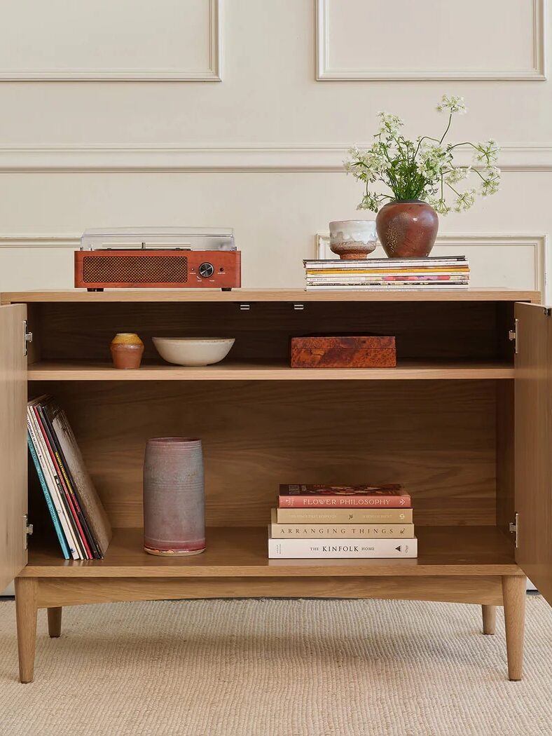 A wooden cabinet with open doors displays books and decor items inside, while a record player, books, and vases are arranged on top. The cabinet stands against a beige paneled wall.