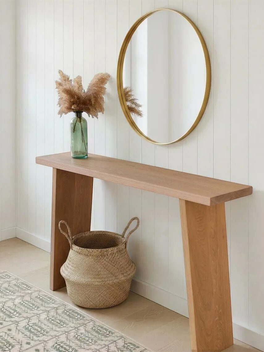 A wooden console table with a glass vase of dried pampas grass, a woven basket underneath, and a round wall mirror above against a white paneled wall.