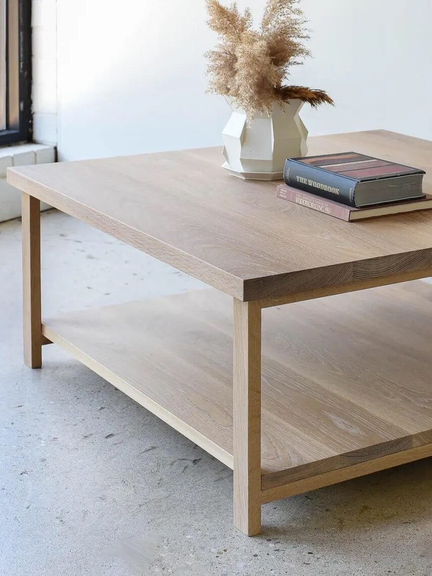 A light wood coffee table with a lower shelf, holding a white vase with dried grass and two stacked books, placed on a concrete floor near a window.