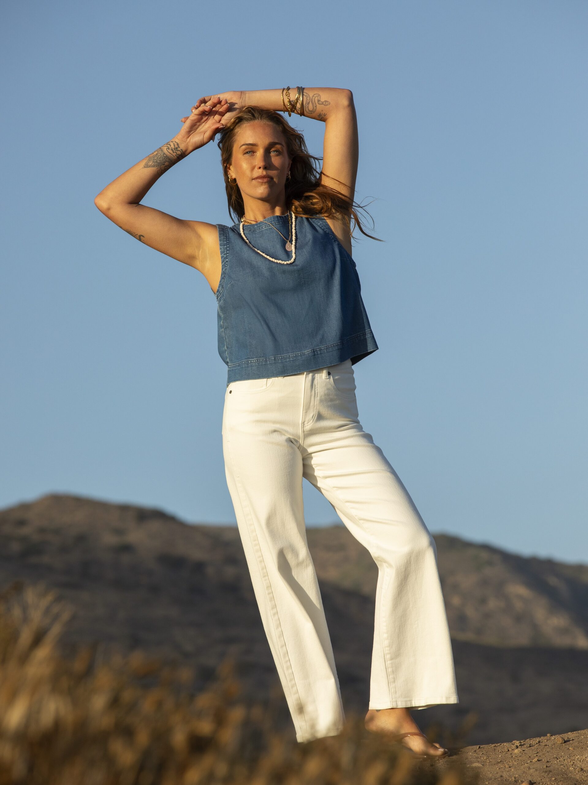 Woman standing outdoors on a grassy hill, wearing a sleeveless blue top and white pants, with arms raised above her head. Mountains and clear blue sky in the background.