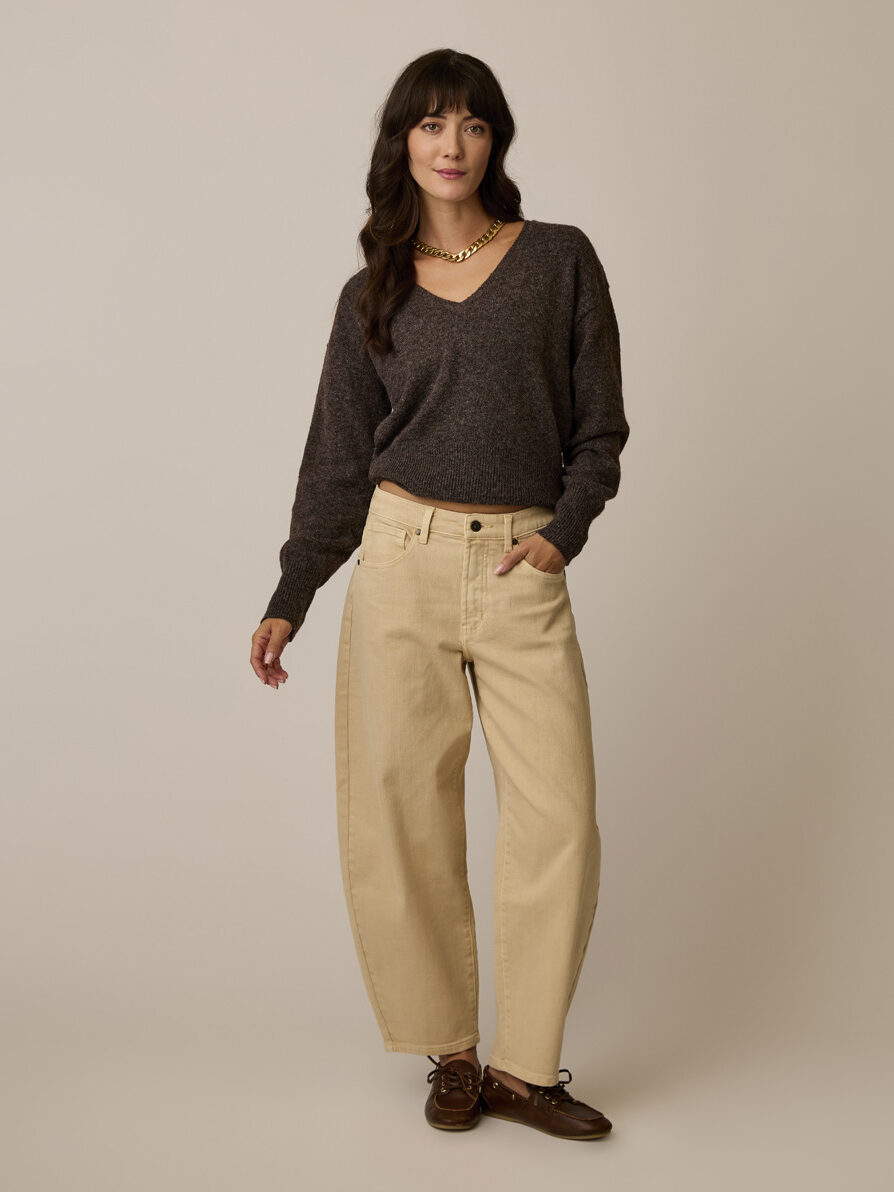 Woman stands against a neutral background wearing a dark V-neck sweater, loose beige jeans, brown shoes, and a chunky gold necklace.