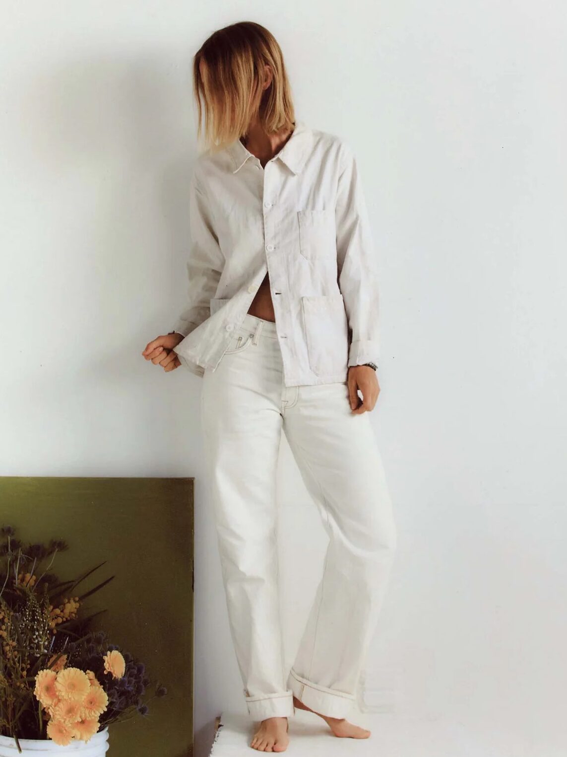 Person standing barefoot against a white wall, wearing a light-colored button-up shirt and loose white pants, with head slightly turned and flowers in a white bucket nearby.