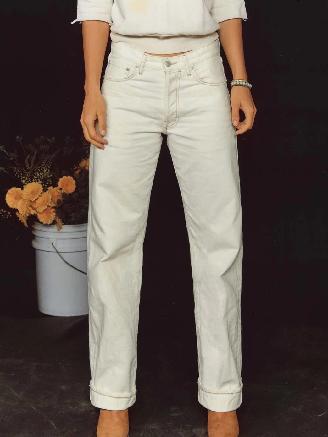 A person wearing white jeans, a white top with rolled sleeves, brown shoes, and a bracelet stands in front of a black background with a bucket of flowers.