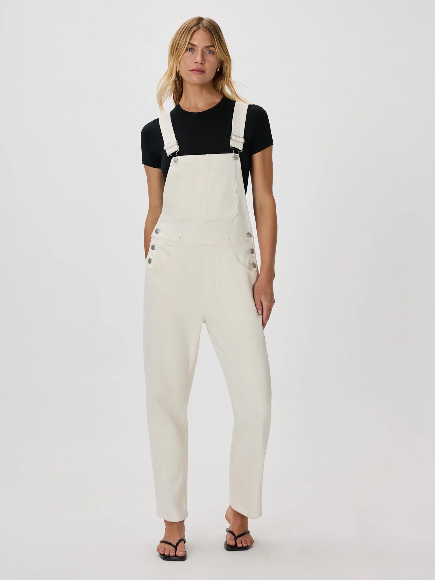 Person stands facing forward wearing a black t-shirt, white overalls, and black sandals against a plain white background.