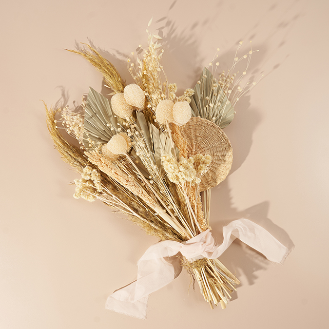 A bouquet of dried flowers and grasses, including palm leaves and round white blooms, tied with a white ribbon, arranged on a neutral beige background.