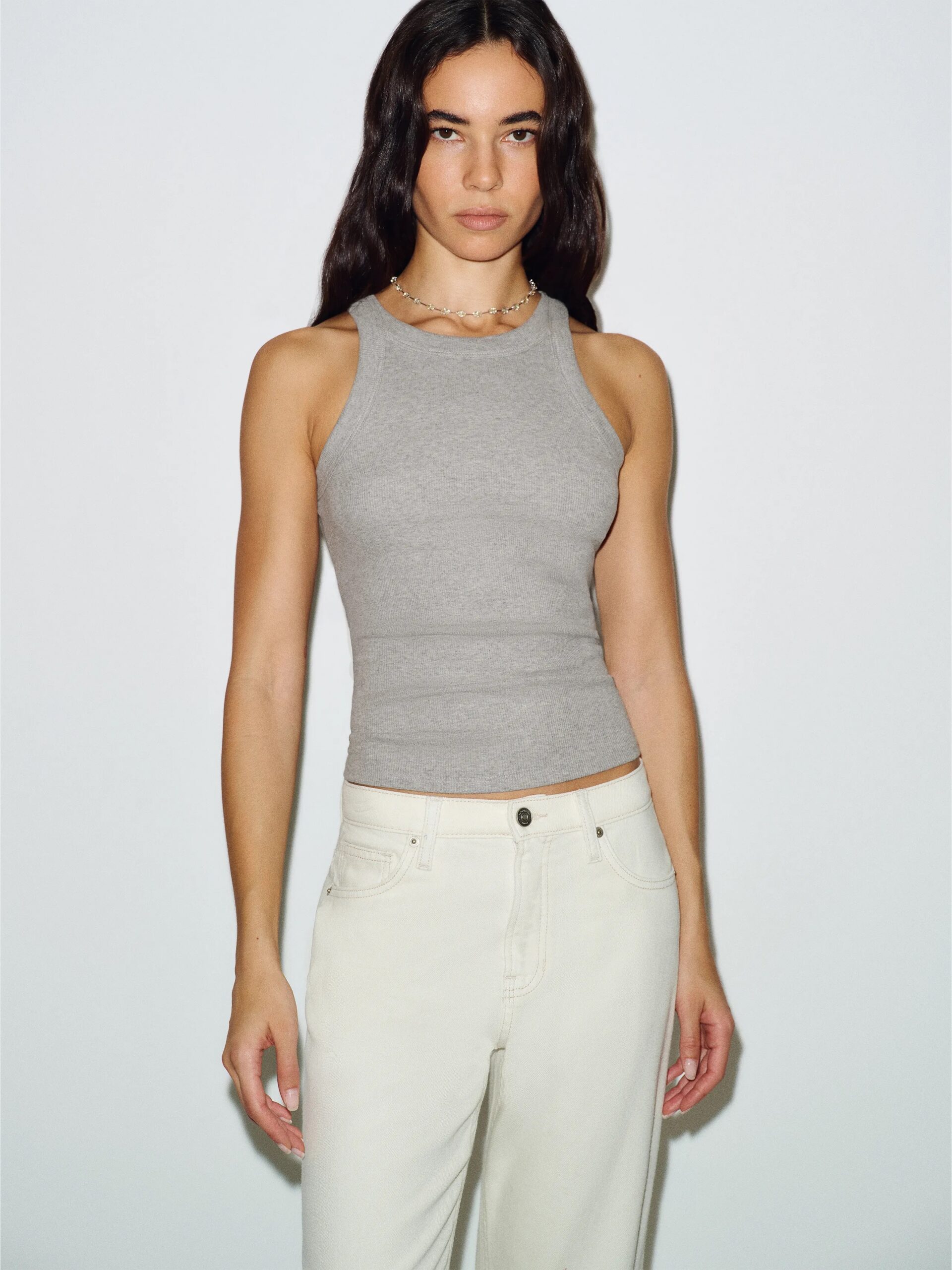 A person with long dark hair wears a gray sleeveless top and light-colored pants, standing against a plain white background.