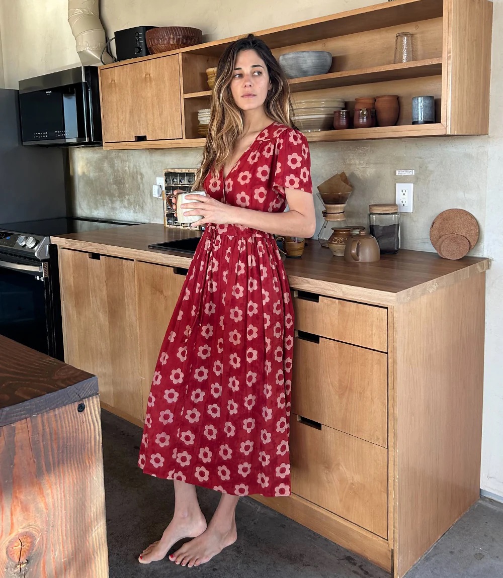 A woman in a red floral dress stands barefoot in a modern kitchen, holding a mug, with wooden cabinets and shelves in the background.