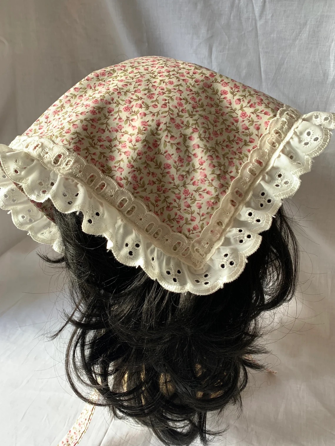 A pink floral kerchief with lace trim is placed on a dark-haired wig, set against a plain white background.