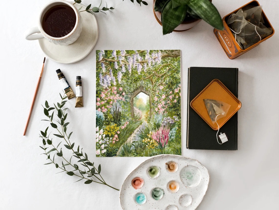 A watercolor painting of a garden scene is surrounded by art supplies, a cup of tea, two potted plants, and an open tea tin on a white surface.