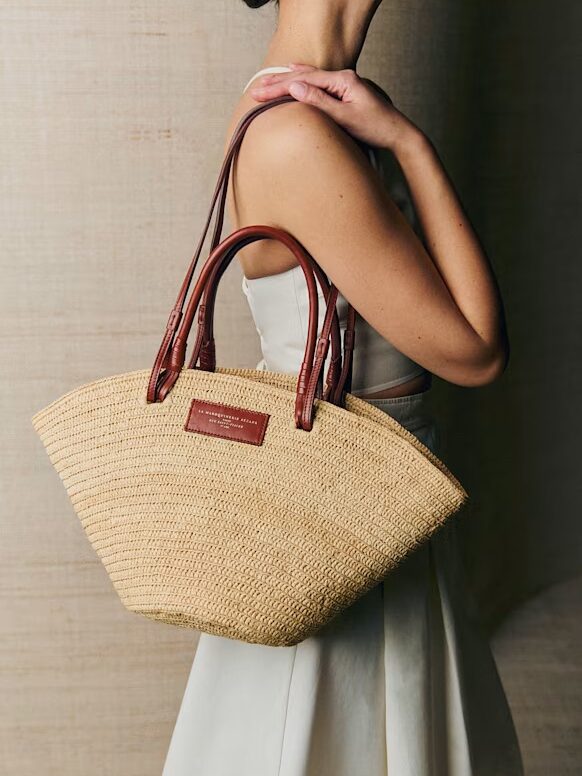 A woman in a white sleeveless dress carries a woven straw handbag with brown leather handles over her shoulder.