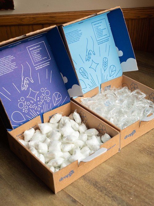 Two open cardboard boxes filled with clear and white detergent pods, with illustrated instructions printed on the inside lids.