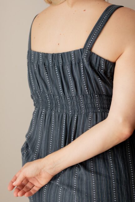 A person in a blue, pinstriped sleeveless dress stands with one arm bent behind their back, showing the back of the dress and part of their shoulder.
