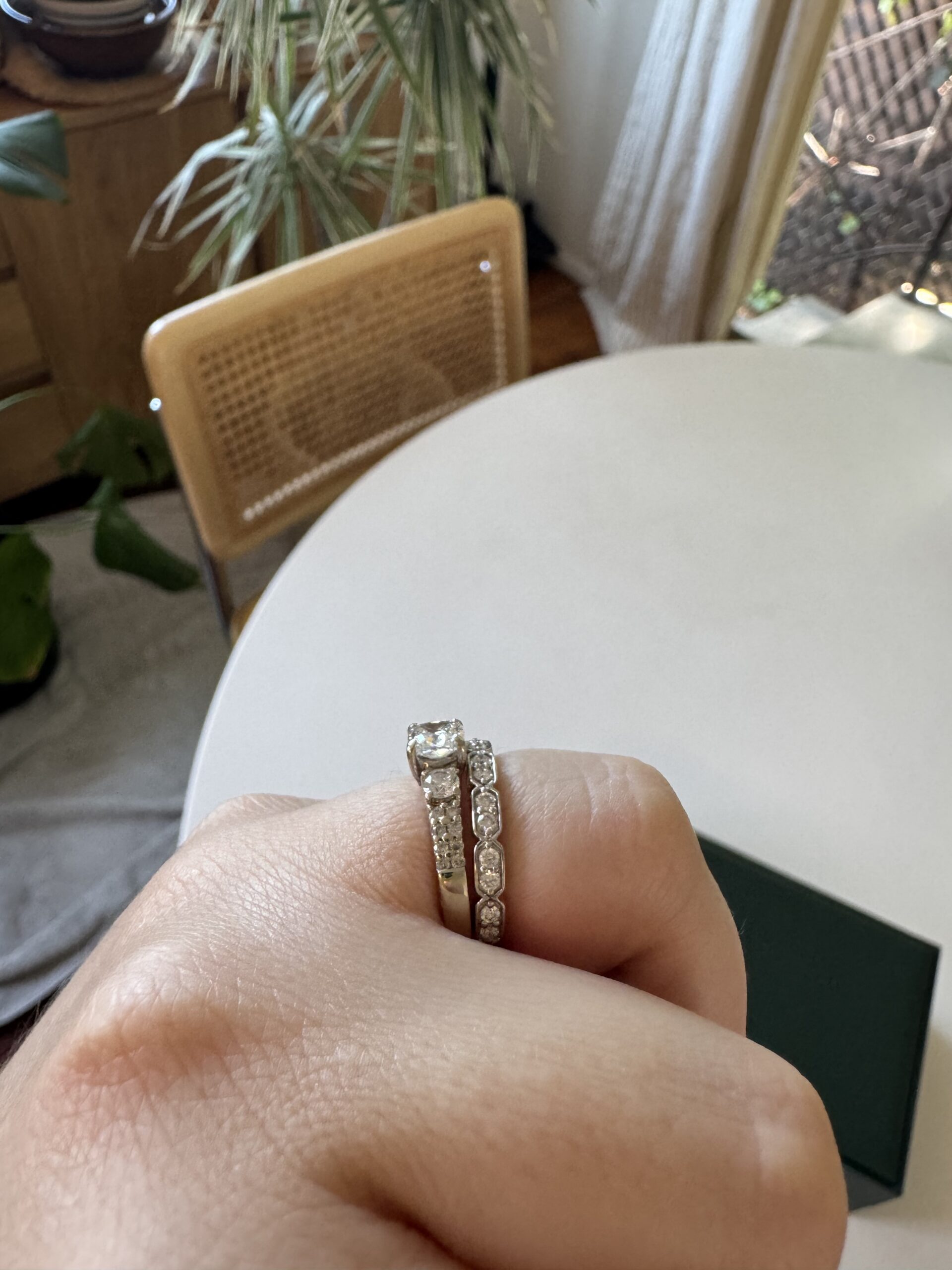 A hand with two diamond rings is held over a white round table, with a wooden chair and indoor plants in the background.