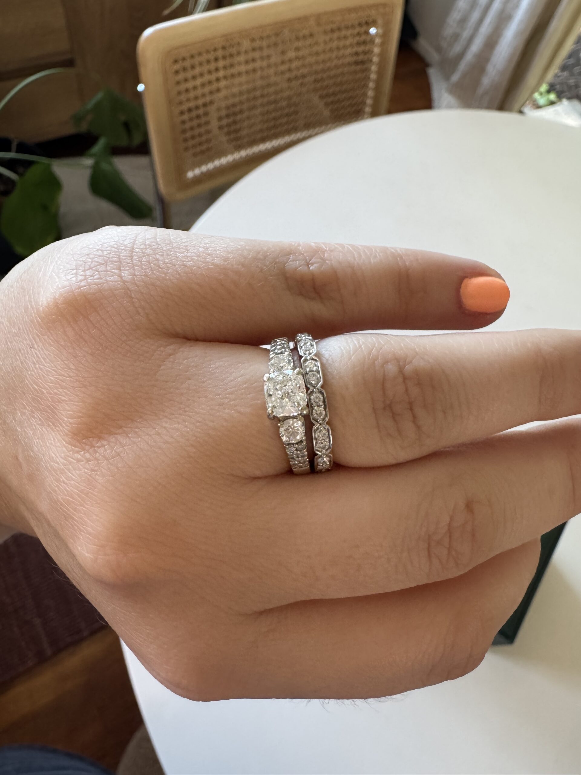 A hand with coral-painted nails wears a solitaire diamond engagement ring stacked with two diamond-studded wedding bands.