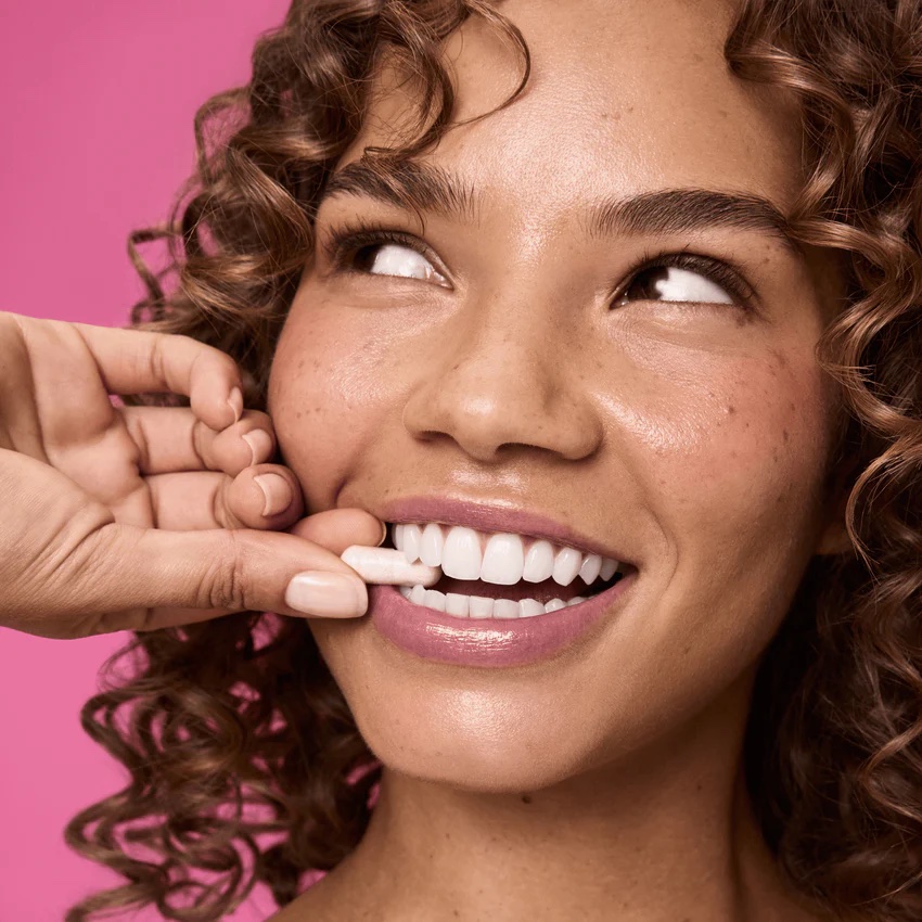 A person with curly hair smiles as another hand holds a white tooth whitening strip against their upper teeth. The background is pink.