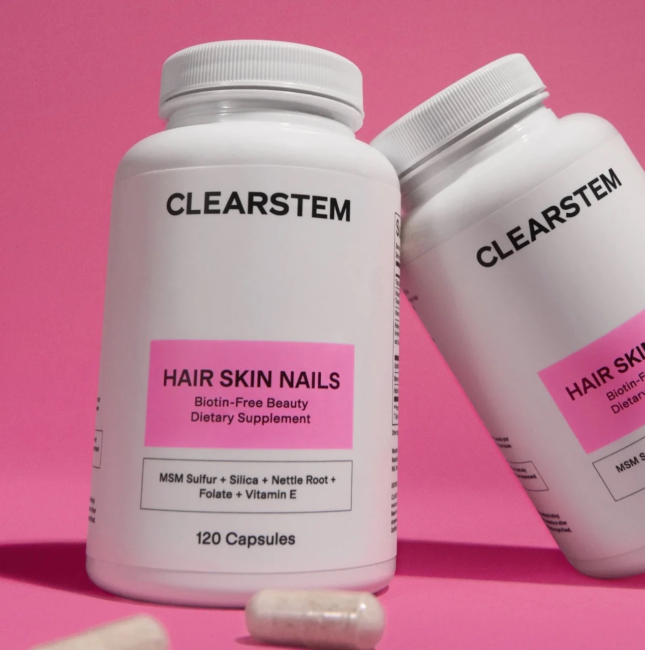 Two white bottles labeled "CLEARSTEM Hair Skin Nails" dietary supplement capsules are shown on a pink background, with two loose capsules in the foreground.