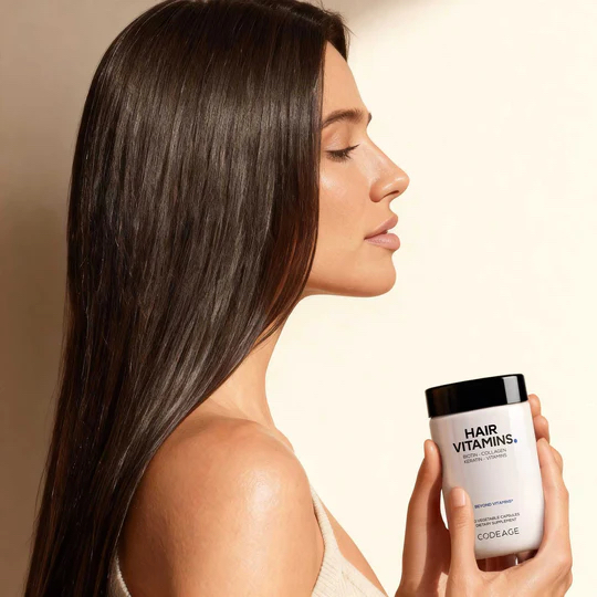 A woman with long straight hair holds a bottle labeled "Hair Vitamins" by Codeage while facing sideways against a neutral background.