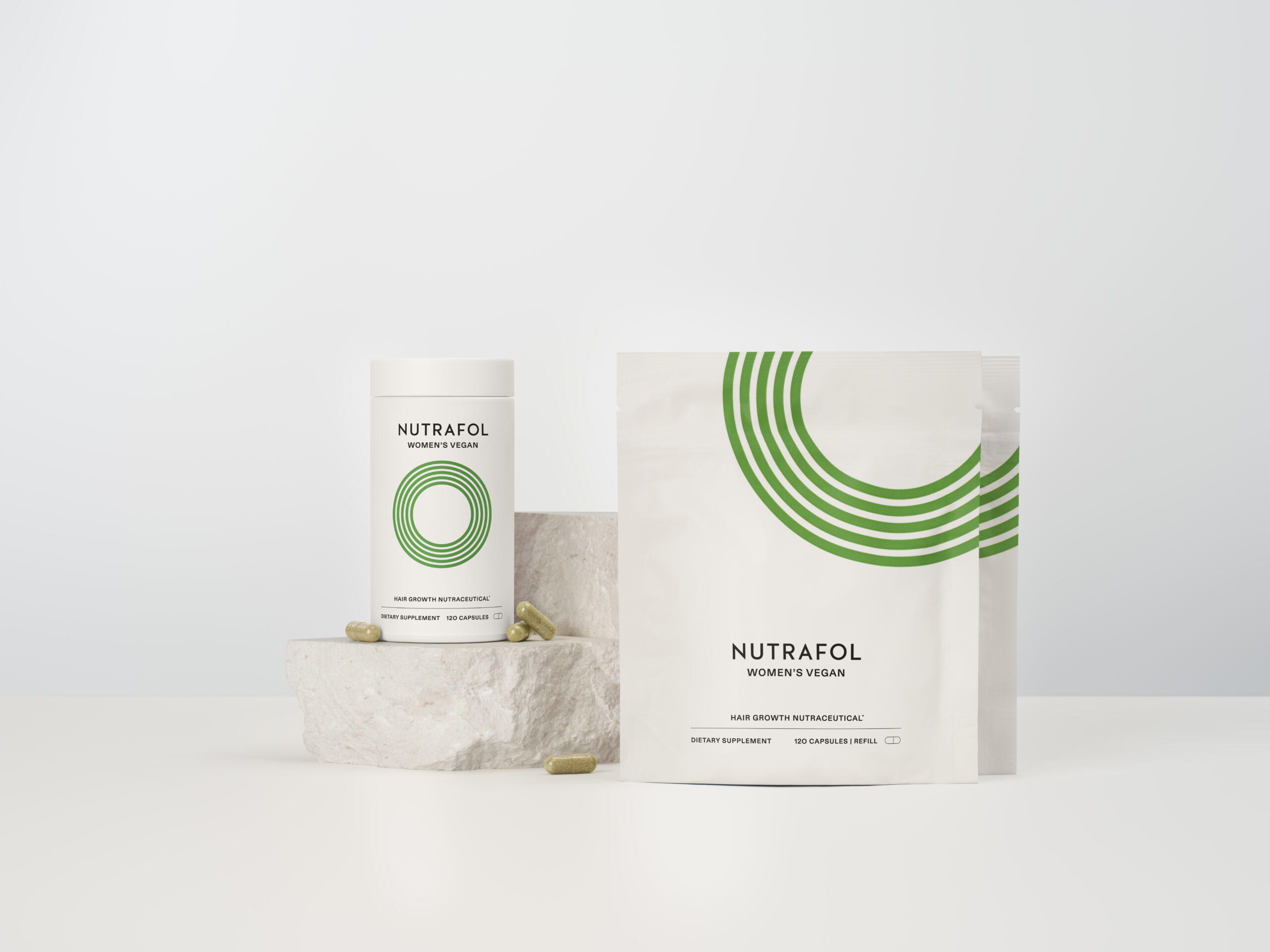 Nutrafol Women’s Vegan supplement bottle and pouch with green circular logo, placed on a white stone against a light background, with capsules visible beside the products.