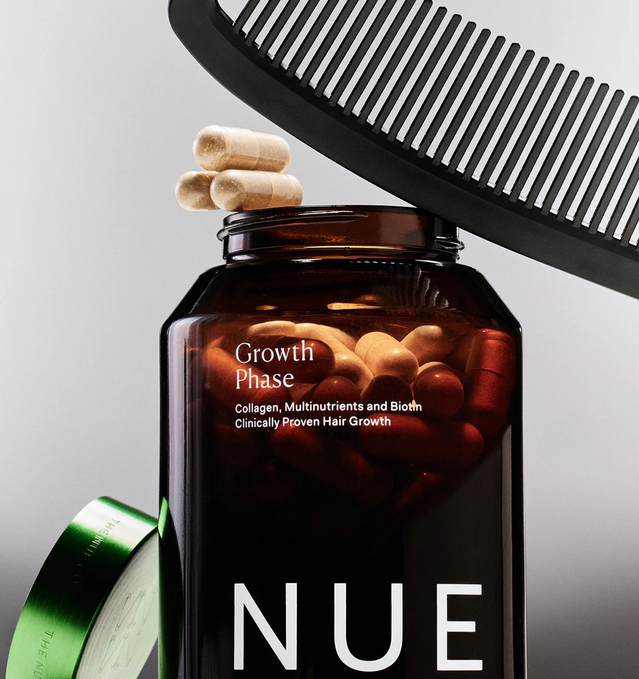 A brown NUE bottle labeled "Growth Phase" with capsules inside and a few on top, a green lid to the side, and a large black comb above the bottle.