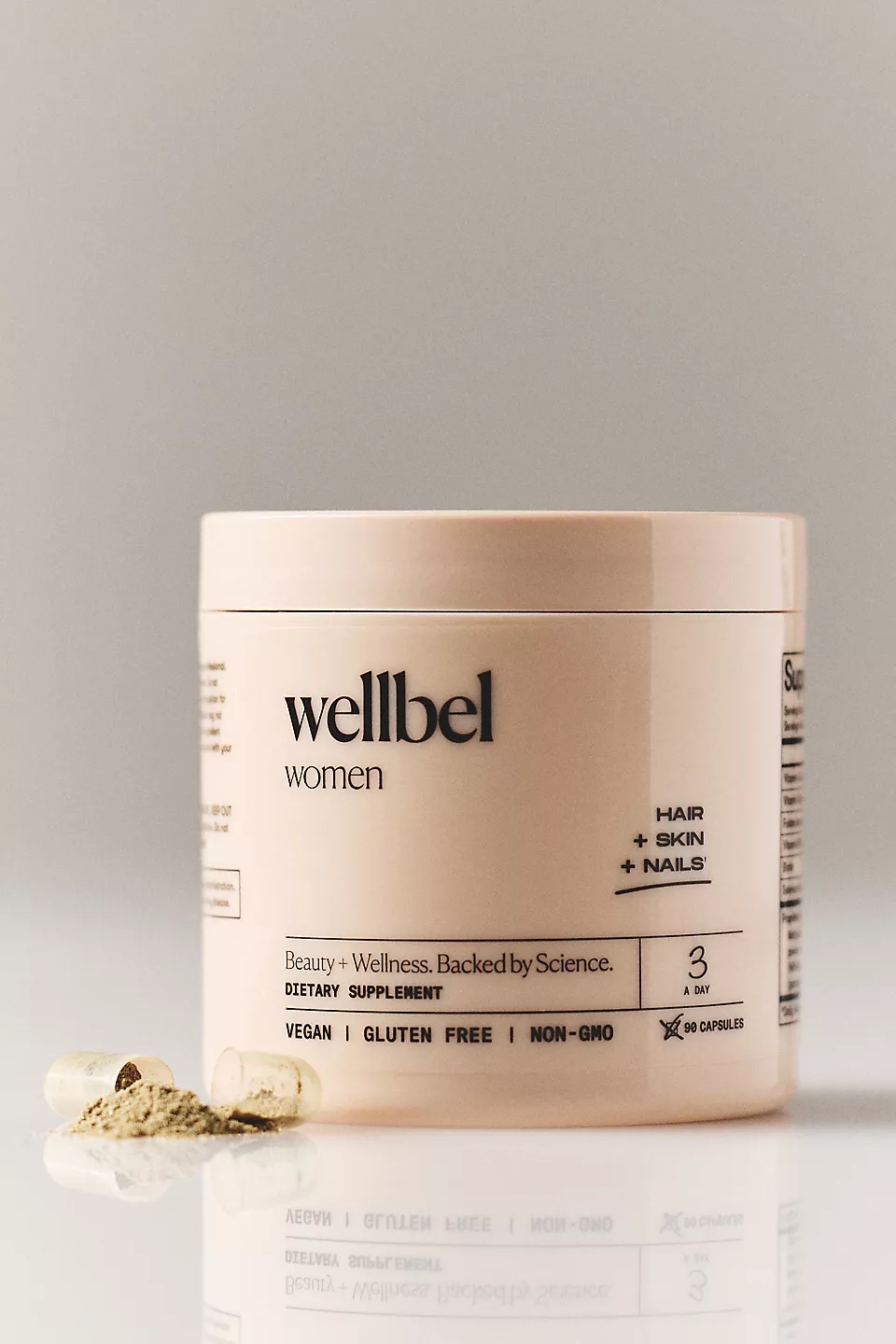 A beige container of Wellbel Women dietary supplement sits on a reflective surface with two capsules and powder beside it. The label highlights benefits for hair, skin, and nails.