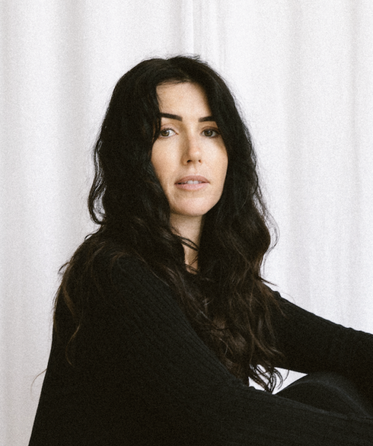 Woman with long dark hair in a black outfit sits barefoot on a white checkered surface against a plain white background, holding a black object in her hand.