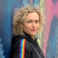 Person with curly blonde hair wearing a black hoodie with a rainbow stripe, standing in front of a colorful, abstract mural.