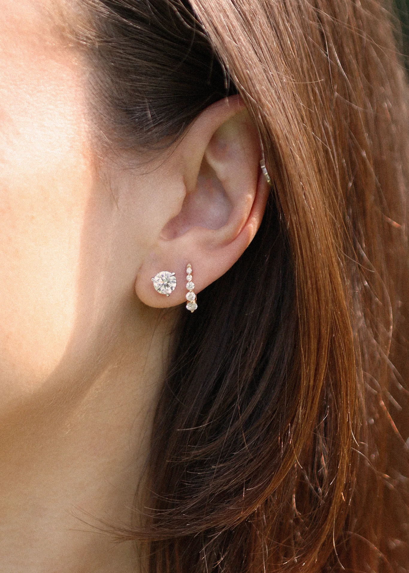 A close-up of a woman’s ear with two earrings: a large round stud and a vertical arrangement of small stones. Brown hair partially covers the ear.