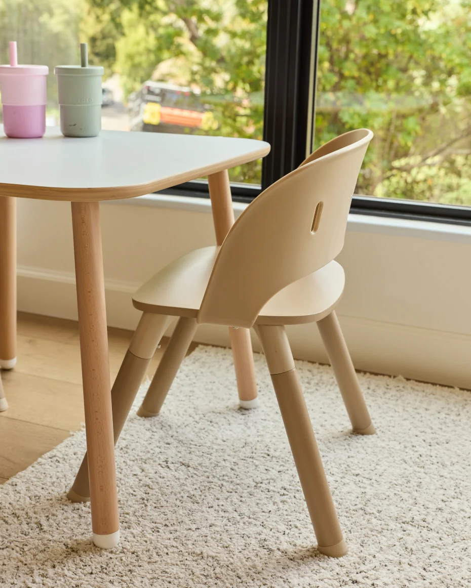 A light-colored wooden children’s chair pulled up to a small table with two cups.