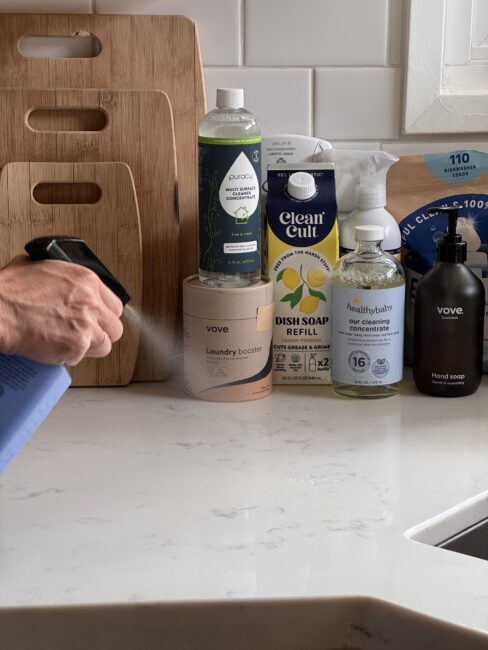A person sprays cleaner onto a white kitchen counter near various cleaning products, bottles, and cutting boards next to a sink.