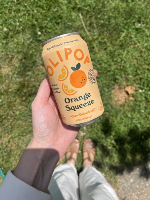 A person holds a can of OLIPOP Orange Squeeze soda outdoors, with grass visible in the background.