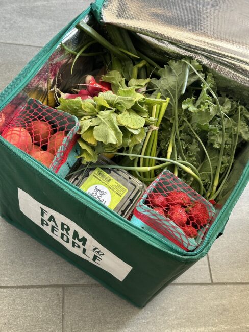 A green insulated bag labeled "Farm to People" filled with fresh leafy greens, radishes, apples, strawberries, and a carton of blueberries.