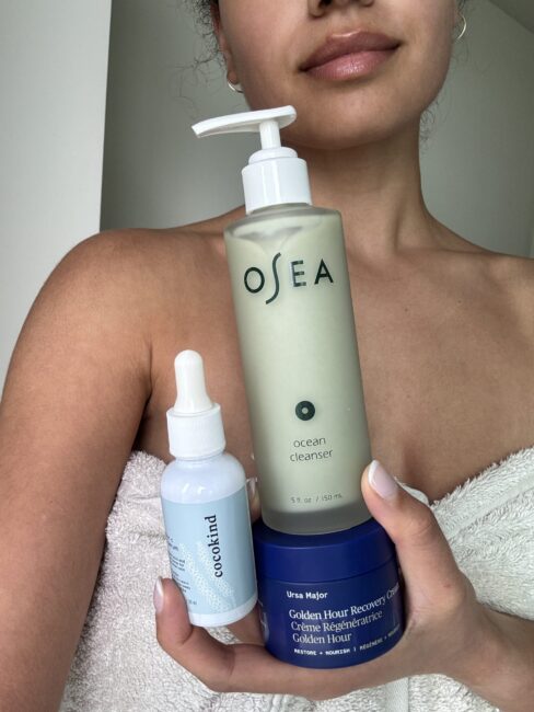 Woman in a towel holds three skincare products: a pump bottle, a blue jar, and a dropper bottle, with minimal background visible.