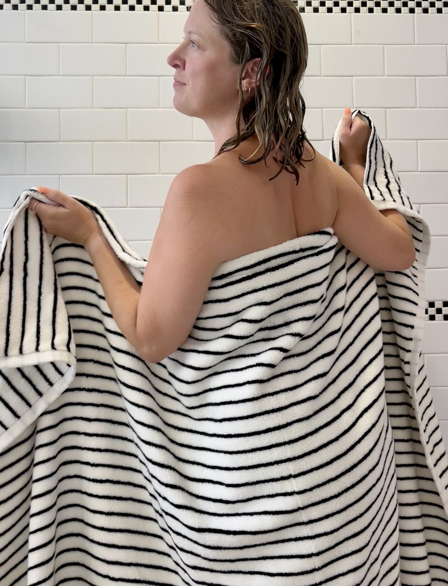 A person with wet hair stands wrapped in a black and white striped organic towel in a tiled bathroom, facing left.