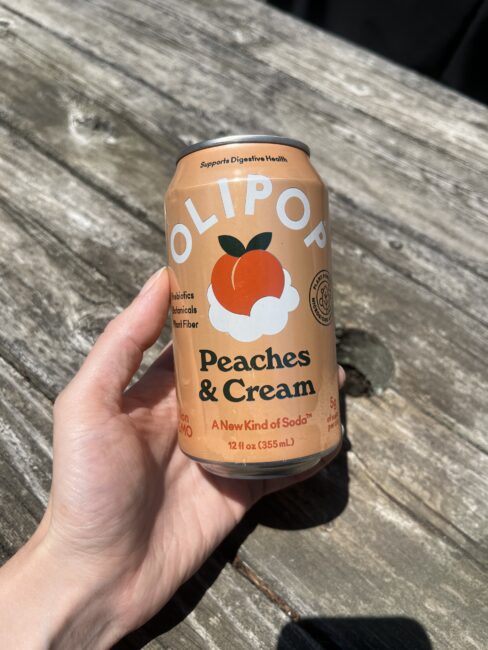 A hand holding a can of OLIPOP Peaches & Cream soda over a weathered wooden surface in sunlight.
