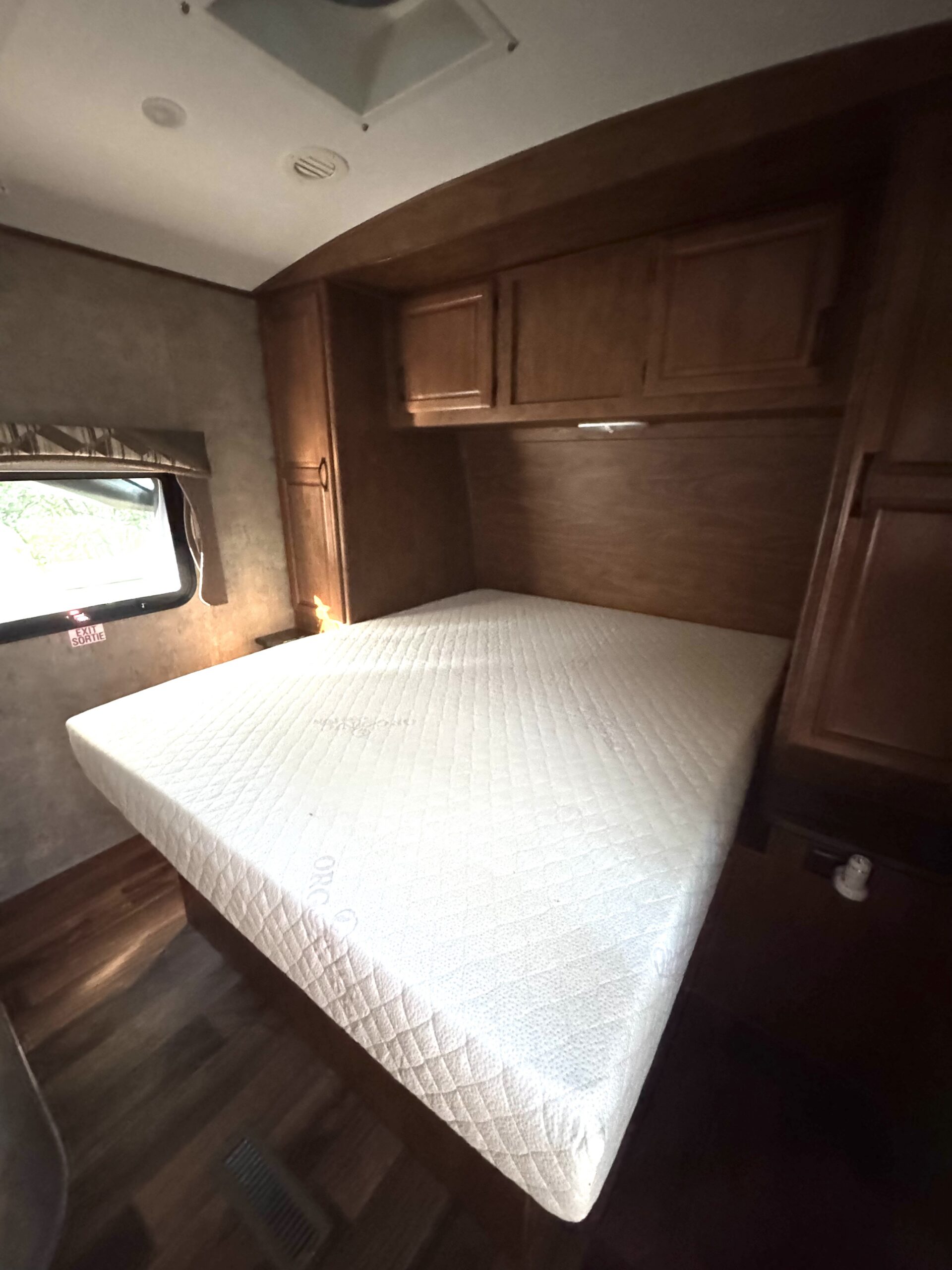 A neatly made bed with a white mattress in a small room with wood-paneled cabinets, a window, and dark wood flooring.